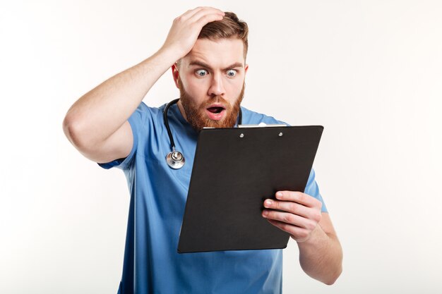 Surprised young doctor holding a clipboard and scratching his head