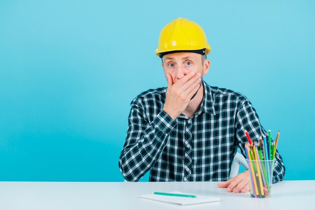 Surprised young engineer is covering his mouth with hand on blue background