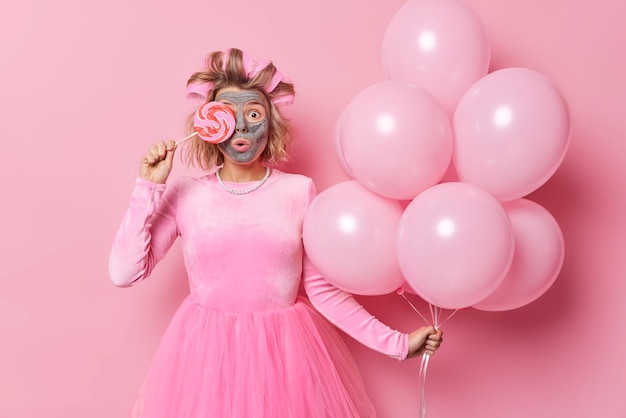 Surprised young European woman covers eye with big caramel candy applies clay facial mask makes hairstyle with rollers wears festive dress holds bunch of balloons going to have birthday party