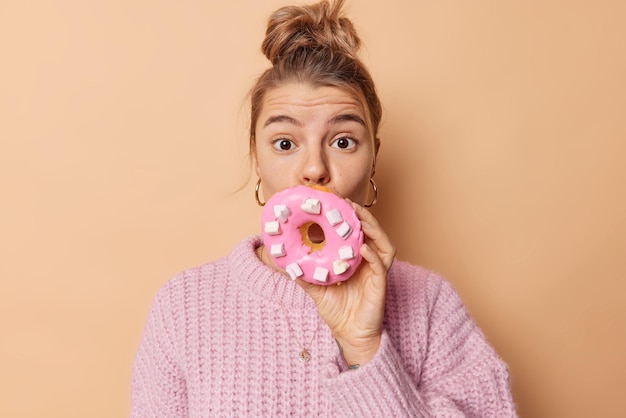 Бесплатное фото Удивленная молодая европейская женщина прикрывает рот вкусным большим аппетитным пончиком, имеет волосы от сахарной зависимости, зачесанные в булочку, носит вязаный свитер, изолированный на бежевом фоне. концепция сладкоежек