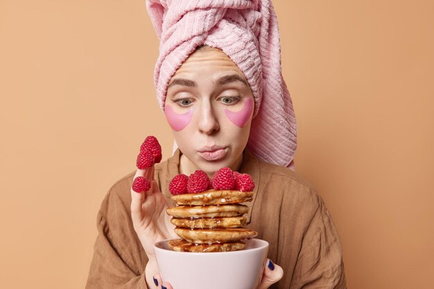 Surprised young European woman looks wondered at appetizing pancakes with syrup and raspberries feels temptation to eat delicious sweet food wears wrapped towel comfortable pajama poses indoor