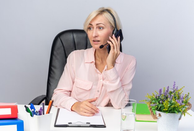 Surprised young female call center operator wearing headset sitting at table with office tools speaks on phone isolated on white wall