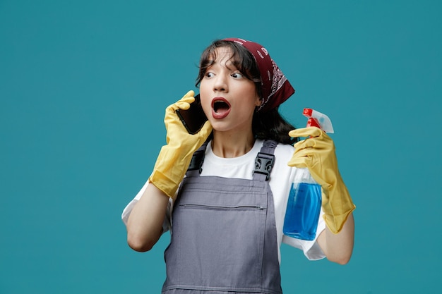 Sorpresa giovane donna addetta alle pulizie che indossa bandana uniforme e guanti di gomma che tengono il detergente guardando di lato parlando al telefono isolato su sfondo blu