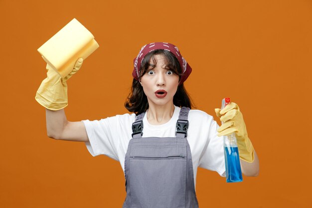 Удивленная молодая женщина-уборщица в униформе резиновых перчаток и бандане поднимает губку, держа моющее средство, глядя на камеру, изолированную на оранжевом фоне