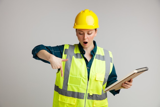 Giovane operaio edile femminile sorpreso che indossa il casco di sicurezza e il blocchetto per appunti della tenuta della maglia di sicurezza che osserva e che indica giù