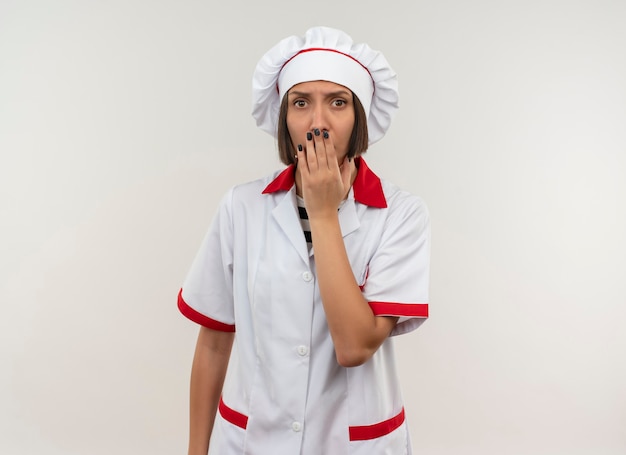 Surprised young female cook in chef uniform putting hand on mouth isolated on white wall