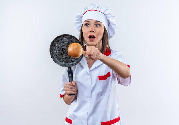 Sorpreso giovane cuoca che indossa l'uniforme dello chef tenendo la padella con il cucchiaio isolato sul muro bianco