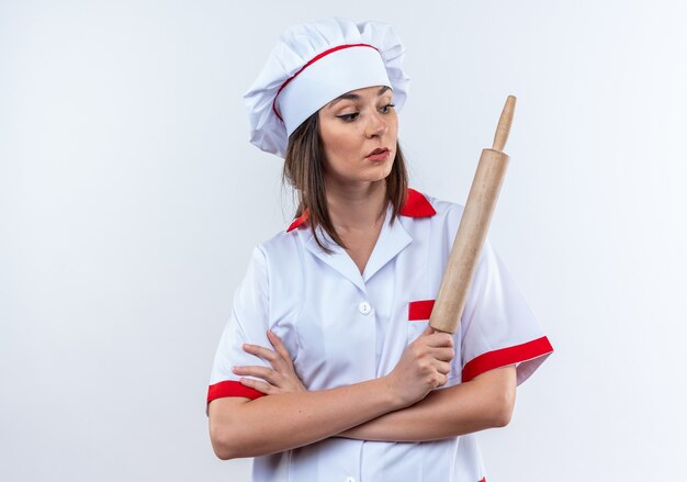 Surprised young female cook wearing chef uniform holding and looking at rolling pin crossing hands isolated on white background