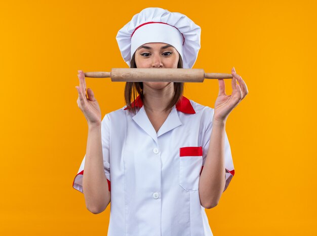 Удивленная молодая женщина-повар в униформе шеф-повара держит и смотрит на скалку, изолированную на оранжевом фоне