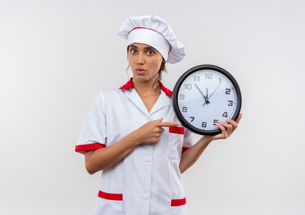 Surprised young female cook wearing chef uniform holding and points to wall clock with copy space