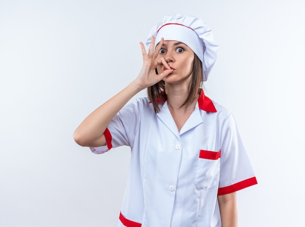удивленная молодая женщина-повар в униформе шеф-повара показывает вкусный жест на белой стене