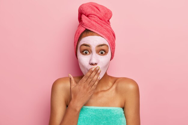 Surprised young female covers mouth with palm, looks at herself in mirror, applies clay facial mask for looking younger and refreshed, stands wrapped in towel, isolated over pink wall