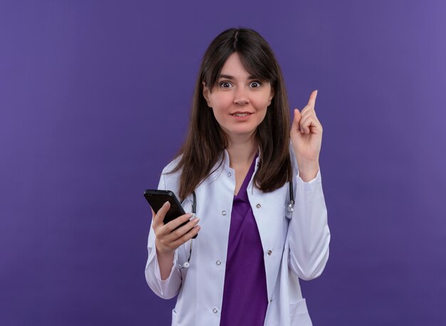 Удивленная молодая женщина-врач в медицинском халате со стетоскопом держит телефон и указывает на изолированный фиолетовый фон с копией пространства