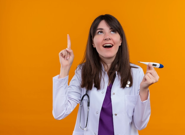 Surprised young female doctor in medical robe with stethoscope holds thermometer and points up on isolated orange background with copy space