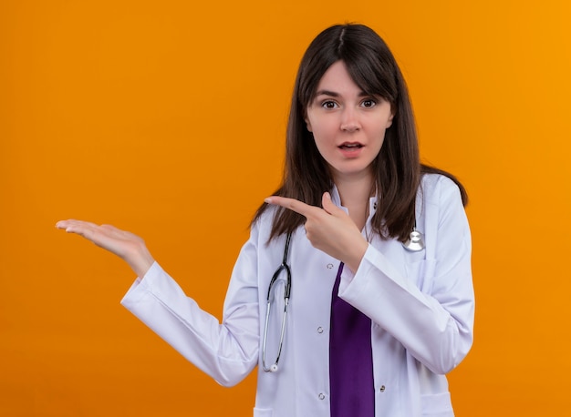 Surprised young female doctor in medical robe with stethoscope points to hand on isolated orange background with copy space