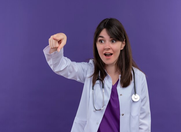 Удивленная молодая женщина-врач в медицинском халате со стетоскопом указывает в сторону на изолированном фиолетовом фоне с копией пространства