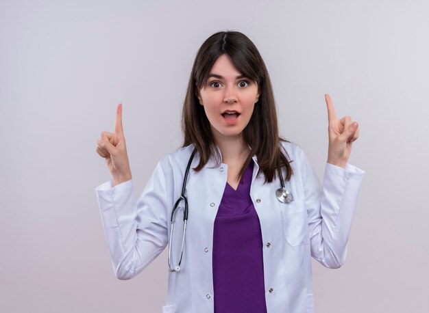 Удивленная молодая женщина-врач в медицинском халате со стетоскопом указывает вверх обеими руками на изолированном белом фоне с копией пространства
