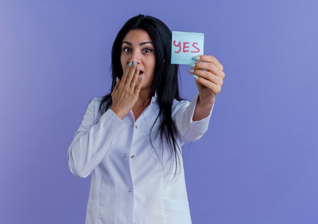 Surprised young female doctor wearing medical robe showing yes note looking keeping hand on mouth isolated