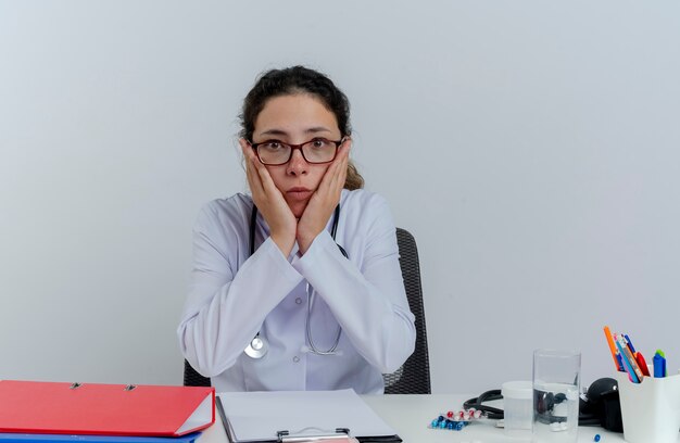 医療用ローブと聴診器と眼鏡を身に着けている驚いた若い女性医師が机に座って医療ツールを見て手を離して顔を隔離している