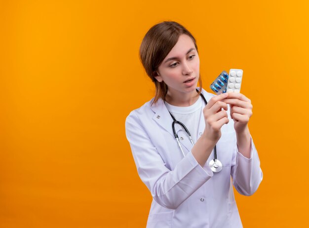 Surprised young female doctor wearing medical robe and stethoscope holding medical drugs and looking at them on isolated orange wall with copy space