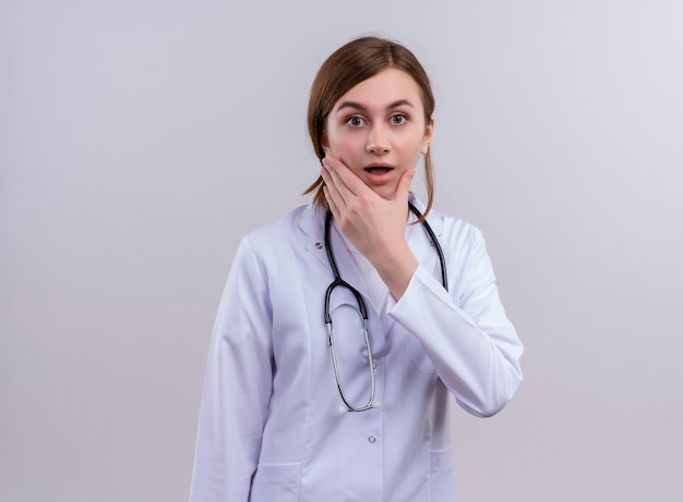 Surprised young female doctor wearing medical robe and stethoscope and putting hand on chin on isolated white wall with copy space