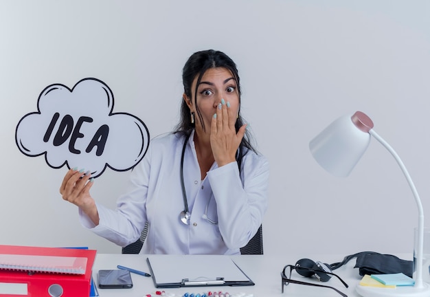 Free photo surprised young female doctor wearing medical robe and stethoscope sitting at desk with medical tools holding idea bubble looking putting hand on mouth isolated