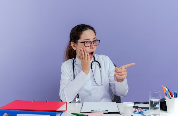 Surprised young female doctor wearing medical robe and stethoscope sitting at desk with medical tools keeping hand on face looking and pointing at side isolated