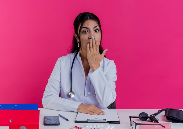 Surprised young female doctor wearing medical robe and stethoscope sitting at desk with medical tools  putting hands on desk and on mouth isolated on pink wall