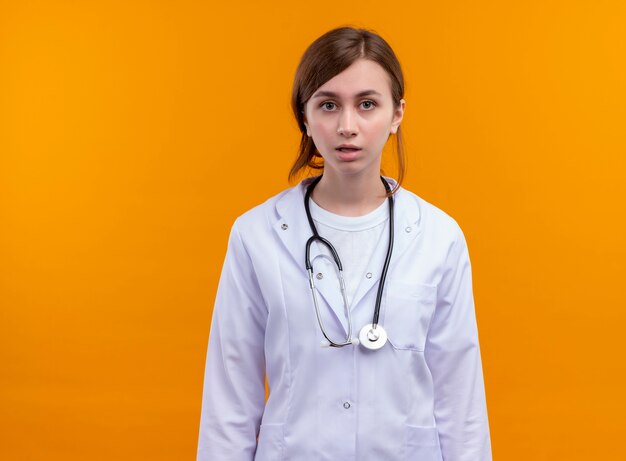 コピースペースのある孤立したオレンジ色の壁に立っている医療ローブと聴診器を身に着けている驚いた若い女性医師