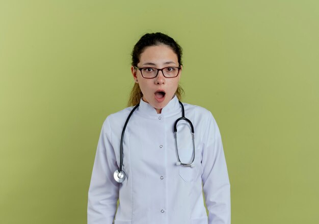 Surprised young female doctor wearing medical robe and stethoscope with glasses isolated on olive green wall