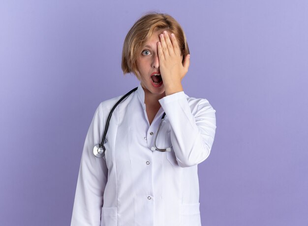 Surprised young female doctor wearing medical robe with stethoscope covered eye with hand isolated on blue background