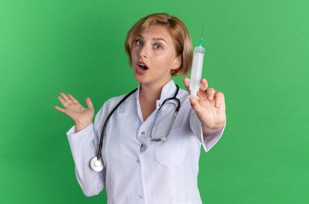 녹색 배경에 격리된 카메라에서 주사기를 들고 있는 청진기가 달린 의료 가운을 입은 놀란 젊은 여성 의사