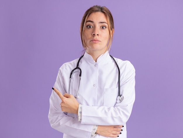 Surprised young female doctor wearing medical robe with stethoscope points at side isolated on purple wall with copy space