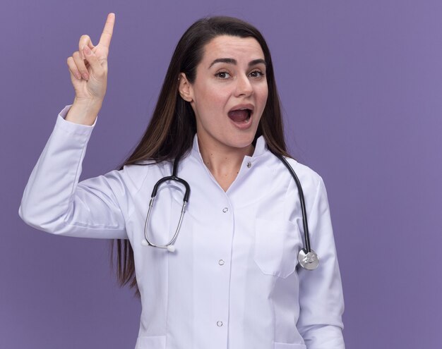 Surprised young female doctor wearing medical robe with stethoscope points up on purple 