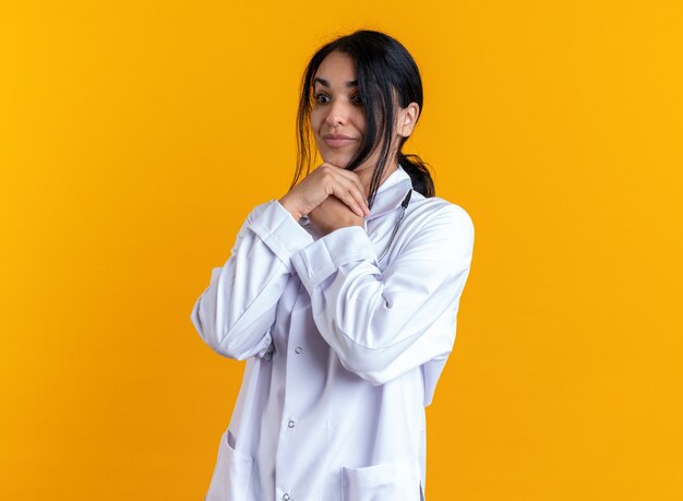 Surprised young female doctor wearing medical robe with stethoscope putting hands under chin isolated on yellow background