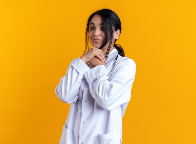 Surprised young female doctor wearing medical robe with stethoscope putting hands under chin isolated on yellow background