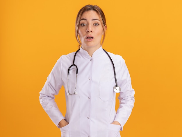 Surprised young female doctor wearing medical robe with stethoscope putting hands in pocket isolated on yellow wall