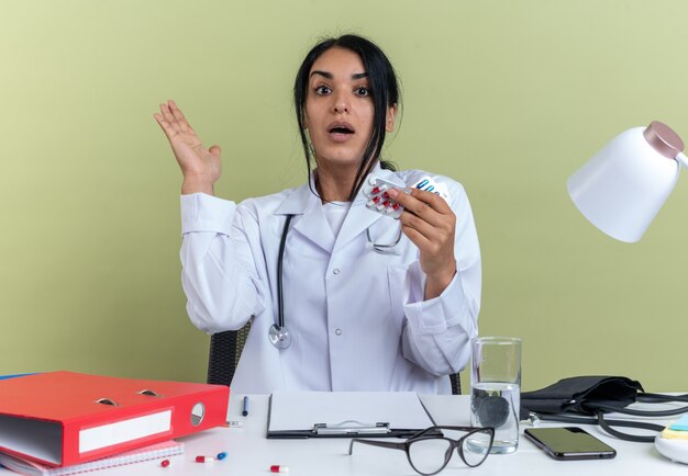 Удивленная молодая женщина-врач в медицинском халате со стетоскопом сидит за столом с медицинскими инструментами, держа таблетки, протягивая руку, изолированную на оливково-зеленом фоне