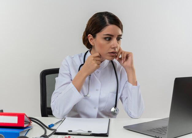 Surprised young female doctor wearing medical robe with stethoscope sitting at desk work on computer with medical tools looking at laptop with copy space