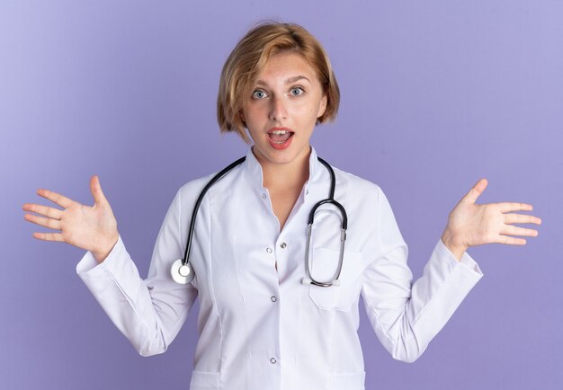 Sorpreso giovane dottoressa che indossa un abito medico con lo stetoscopio che allarga le mani isolate su sfondo blu