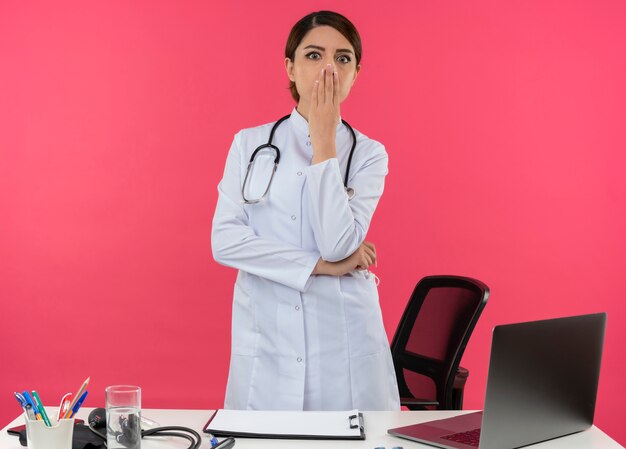 Surprised young female doctor wearing medical robe with stethoscope standing behind desk work on computer with medical tools covered mouth with copy space