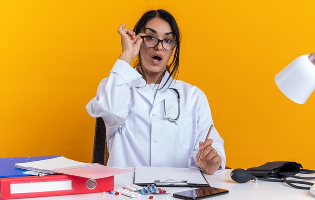 Удивленная молодая женщина-врач в медицинском халате со стетоскопом в очках сидит за столом с медицинскими инструментами, держа карандаш на желтом фоне