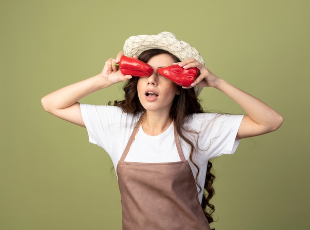 園芸帽子をかぶった制服を着た驚いた若い女性の庭師は、オリーブグリーンの壁に分離された赤唐辛子で目を覆います