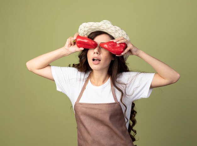 Surprised young female gardener in uniform wearing gardening hat covers eyes with red peppers isolated on olive green wall