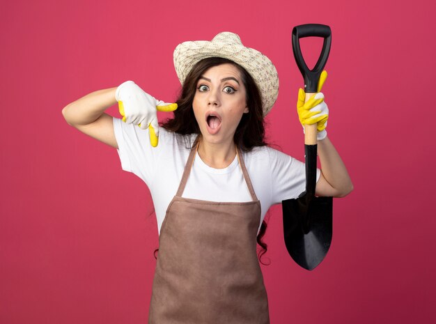 Surprised young female gardener in uniform wearing gardening hat and gloves holds spade and points down isolated on pink wall