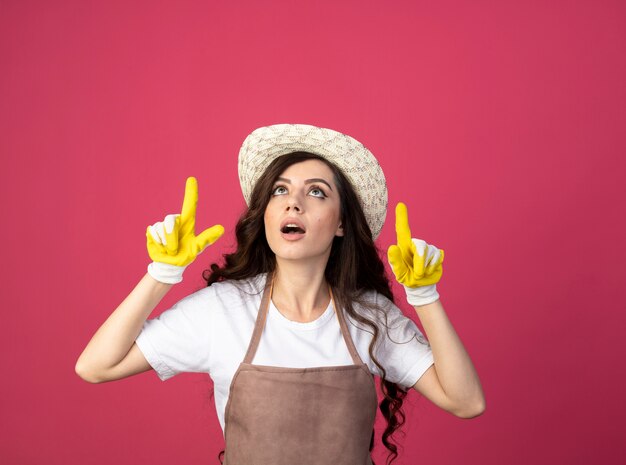 Удивленная молодая женщина-садовник в униформе в садовой шляпе и перчатках смотрит и указывает вверх изолированно на розовой стене