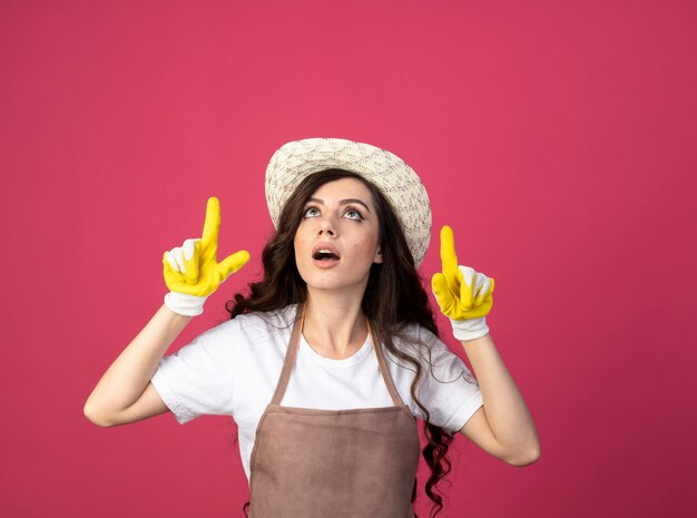 Surprised young female gardener in uniform wearing gardening hat and gloves looks and points up isolated on pink wall