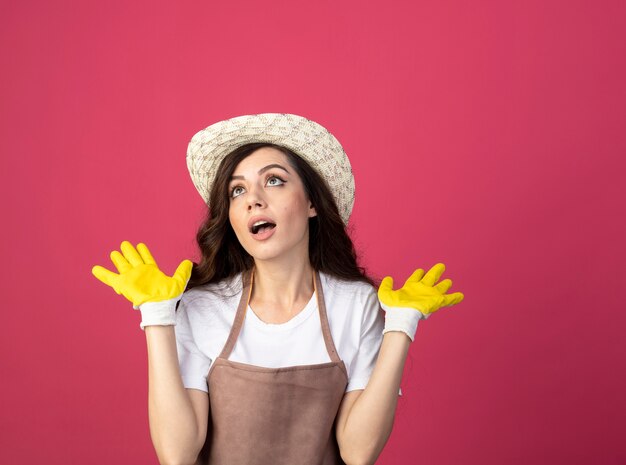 Surprised young female gardener in uniform wearing gardening hat and gloves stands with raised hands looking up isolated on pink wall