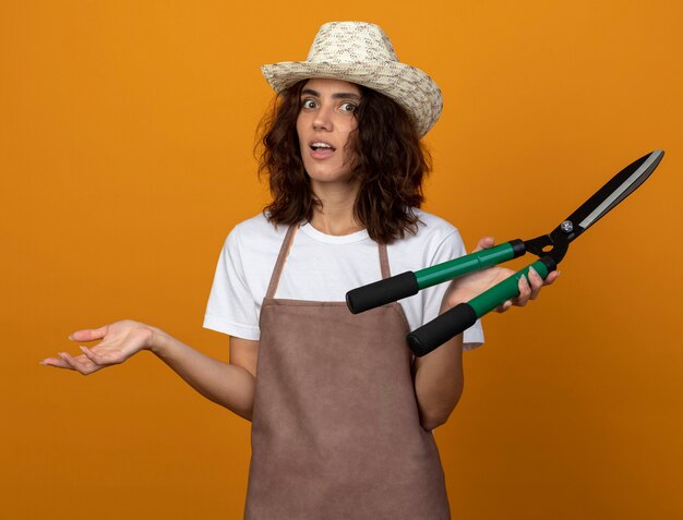 Surprised young female gardener in uniform wearing gardening hat holding clippers and spreading hand