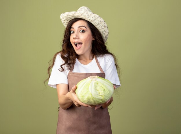 Surprised young female gardener in uniform wearing gardening hat holds cabbage isolated on olive green wall